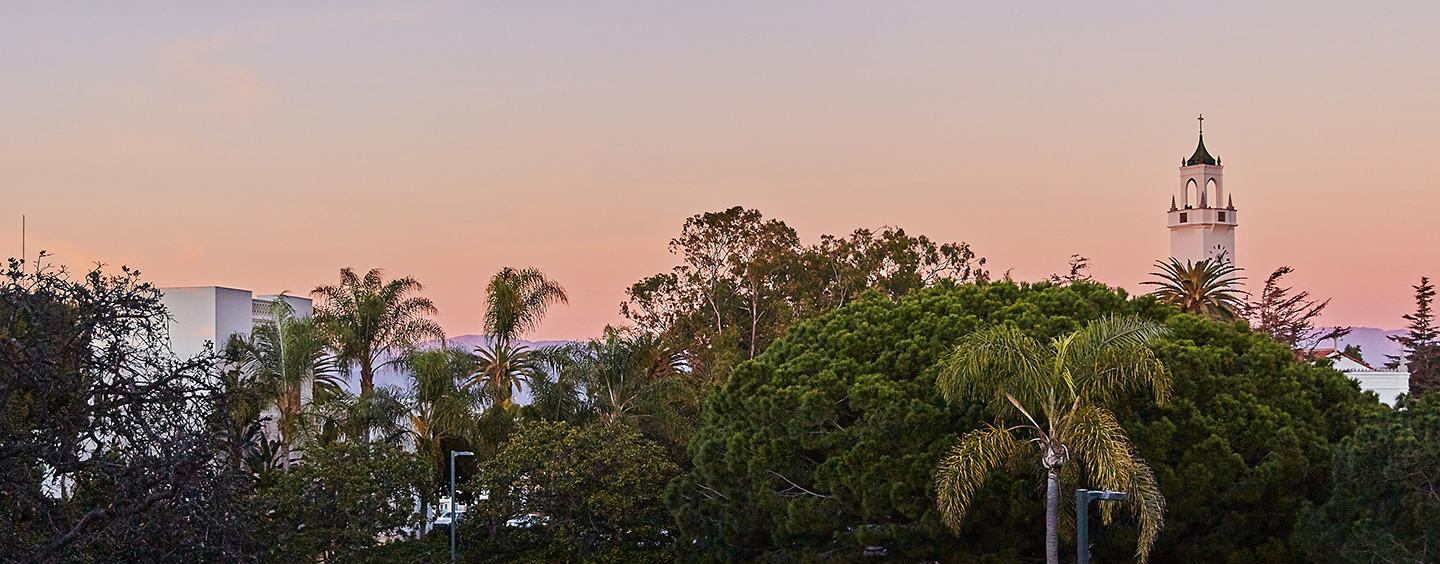 The LMU clock tower at a distance with a pink and purple sunset; lots of greenery in the foreground.